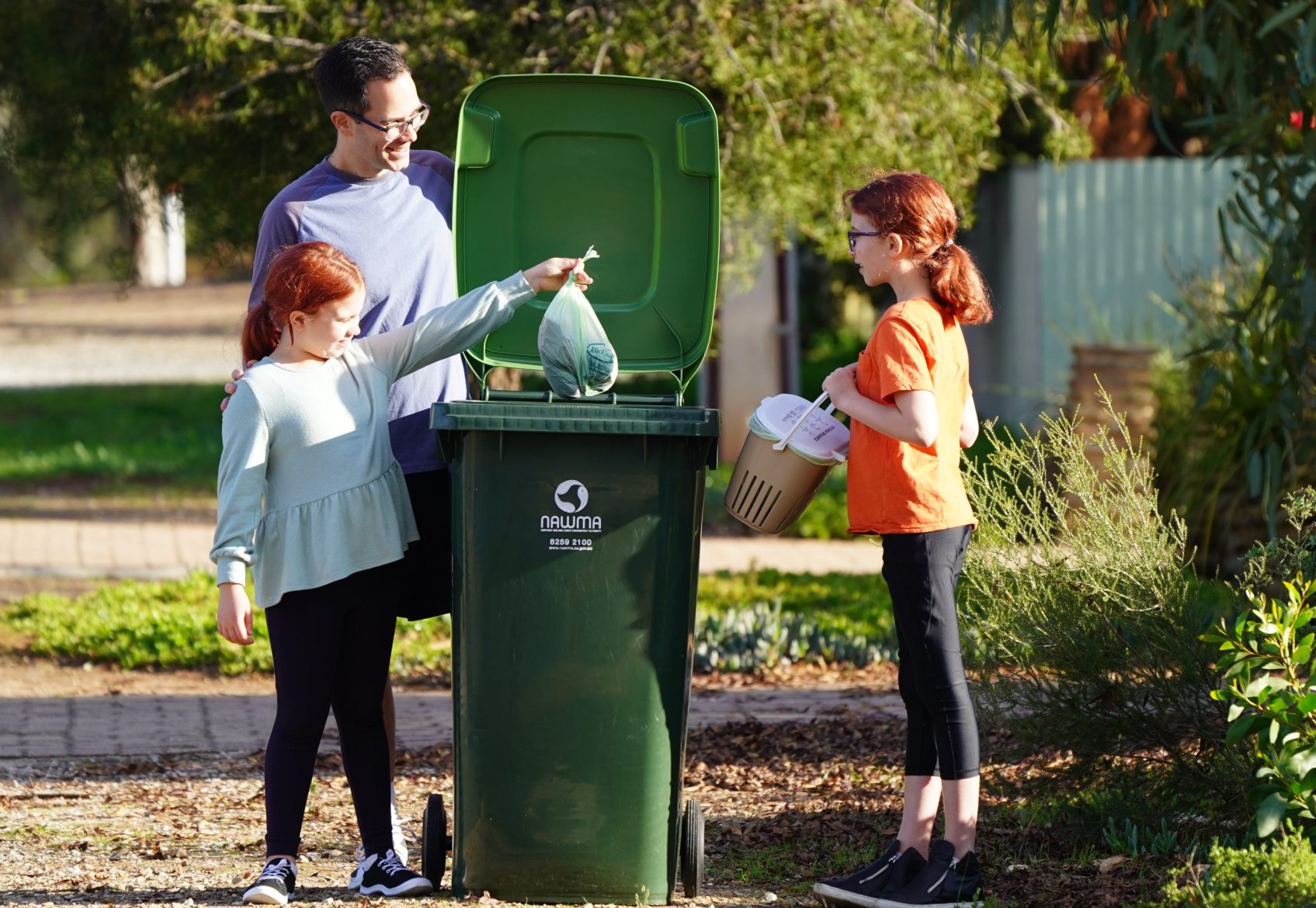 Father standing near kerbside FOGO bin with two children. The father is holding the lid to the bin open and one child is dropping a compostable bag with food scraps into the bin. The other child is holding a ventilated kitchen caddy lined with a compostable bag.